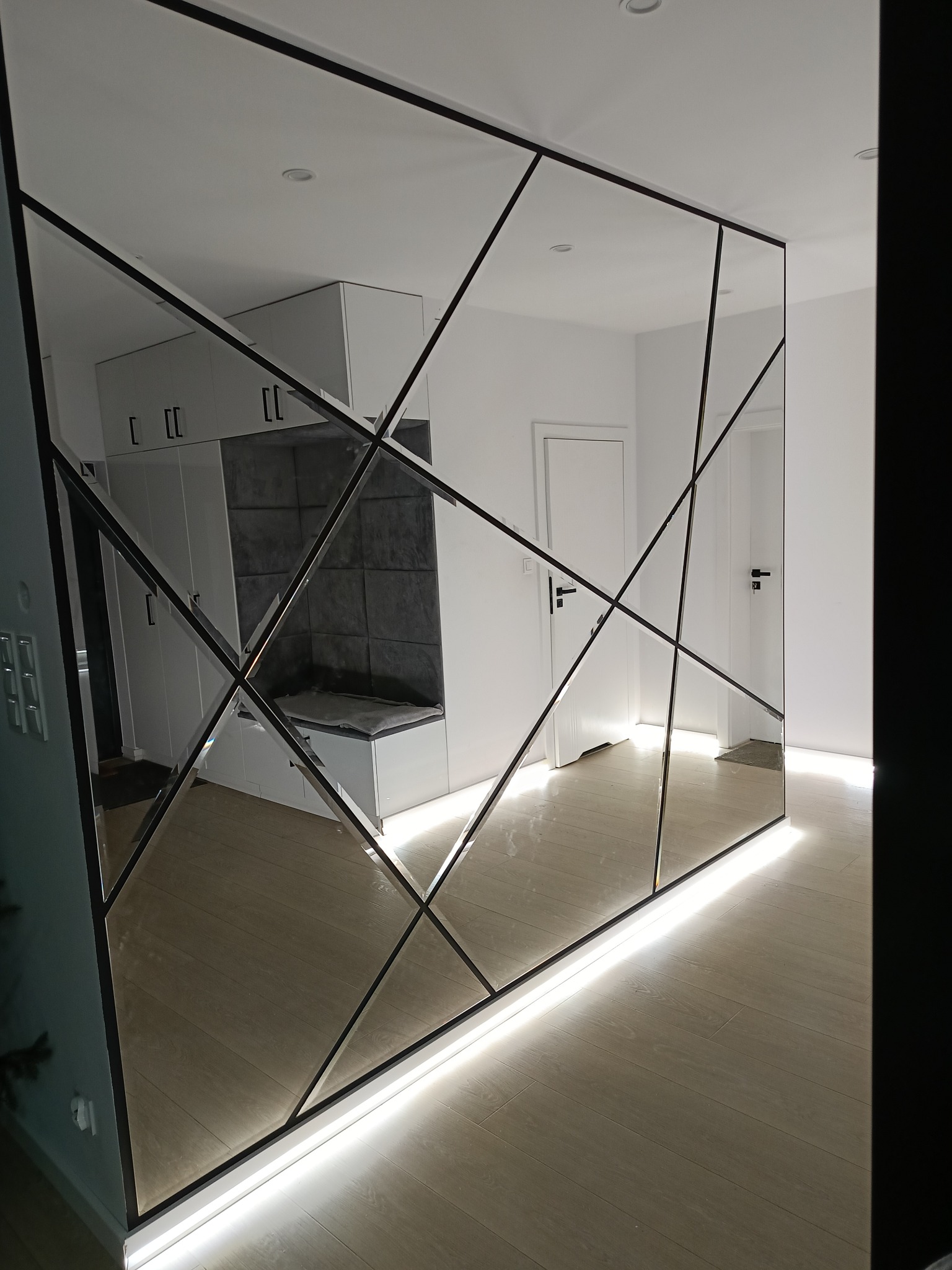 Dekoracyjna ściana lustrzana z podświetleniem LED w Pękaninie. Geometryczny wzór z czarnymi ramami odbija wnętrze: białe szafki i drzwi.