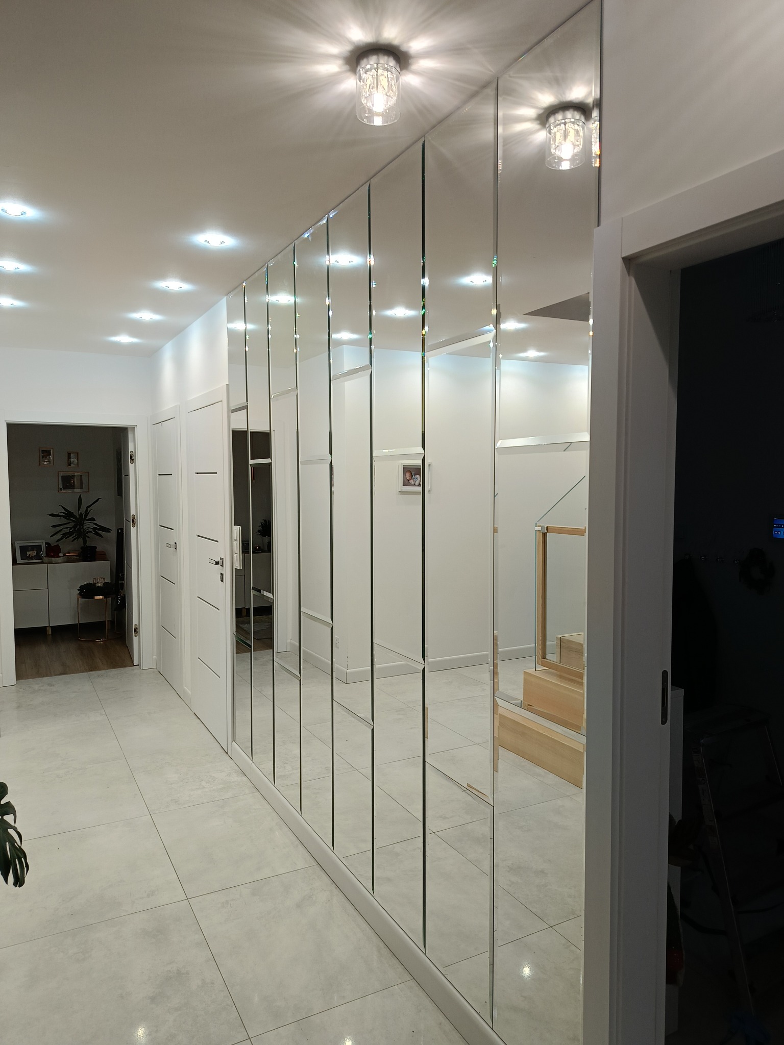 Nowoczesna ściana z luster w przedpokoju, odbijająca schody i wnętrze. Lustra w wąskich panelach z delikatnymi fazowaniami, oświetlenie sufitowe.