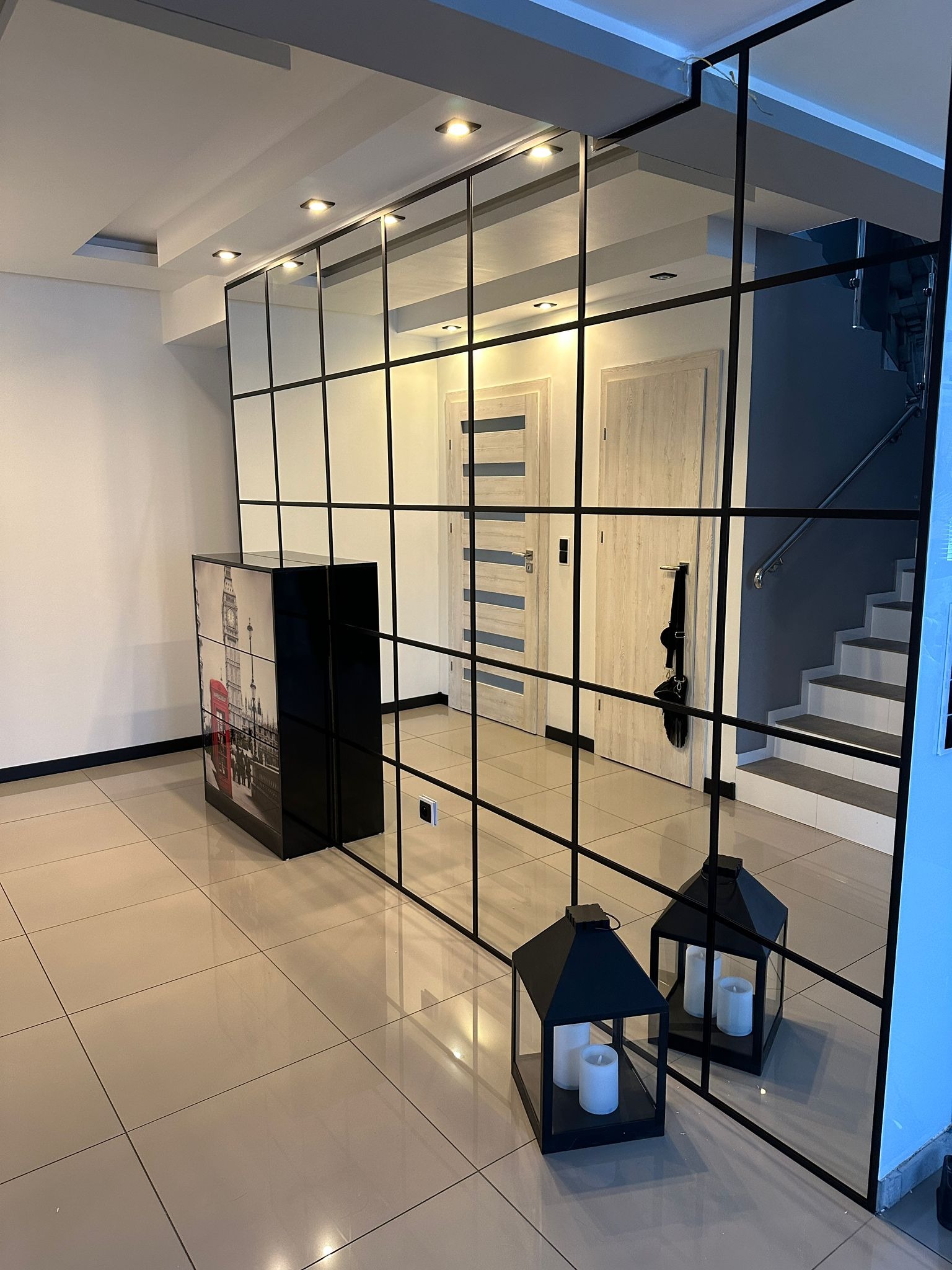 Nowoczesna szklana ścianka działowa z czarnymi profilami, dzieląca przestrzeń w jasnym wnętrzu. Widoczne drzwi i schody w tle, lampiony i szafka z motywem Londynu.