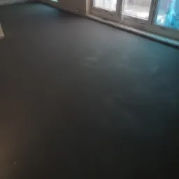 Gładka, ciemnoszara powierzchnia świeżo wylanej wylewki w słabo oświetlonym pomieszczeniu z oknami zasłoniętymi folią.