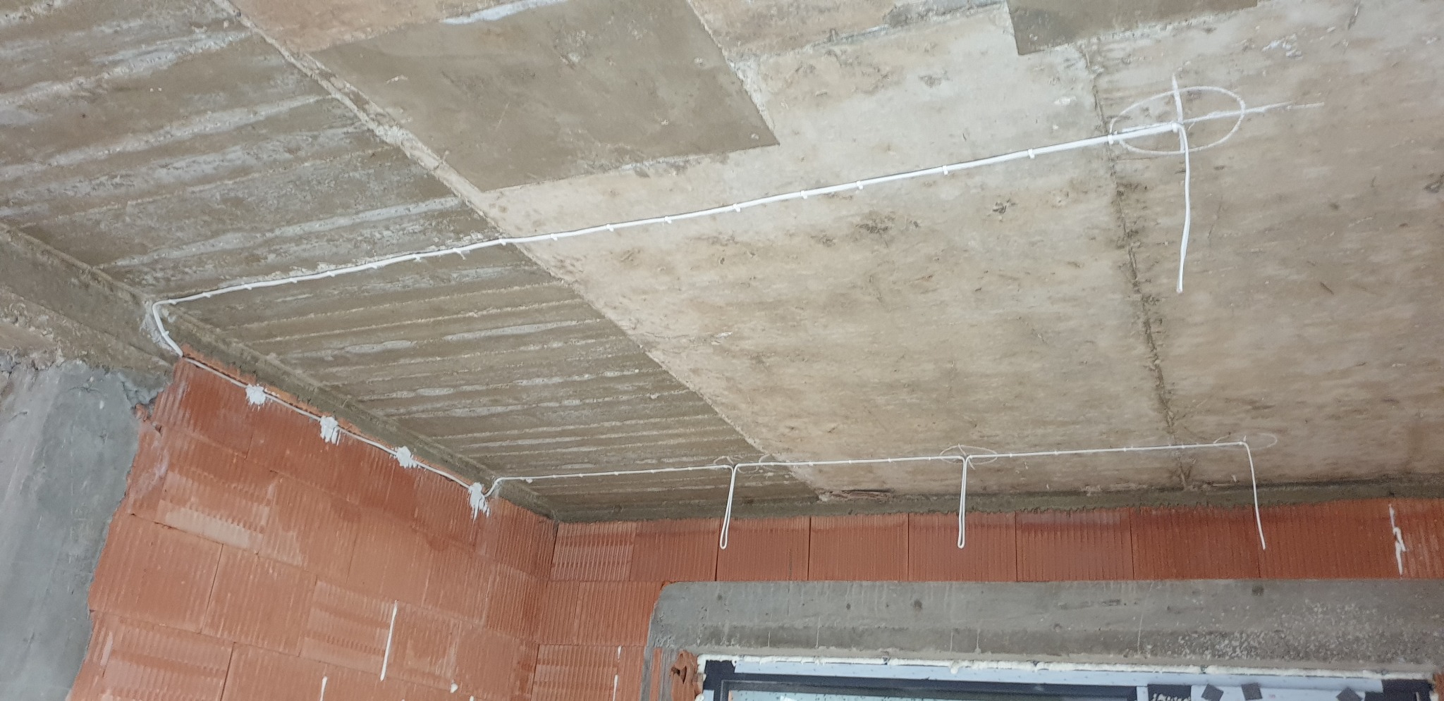 Surowy betonowy strop z widocznymi kablami elektrycznymi prowadzonymi po powierzchni, przytwierdzonymi do ścian z czerwonej cegły. Widoczne wystające końcówki kabli przygotowane do podłączenia...