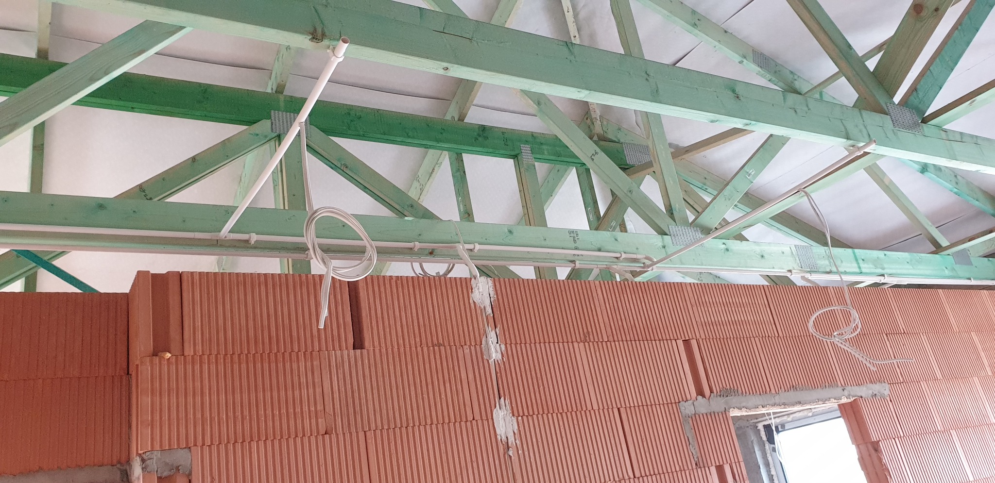 Wewnętrzne okablowanie elektryczne w budynku w stanie surowym: przewody prowadzone pod zielonymi belkami stropowymi, nad czerwoną ścianą z cegieł.