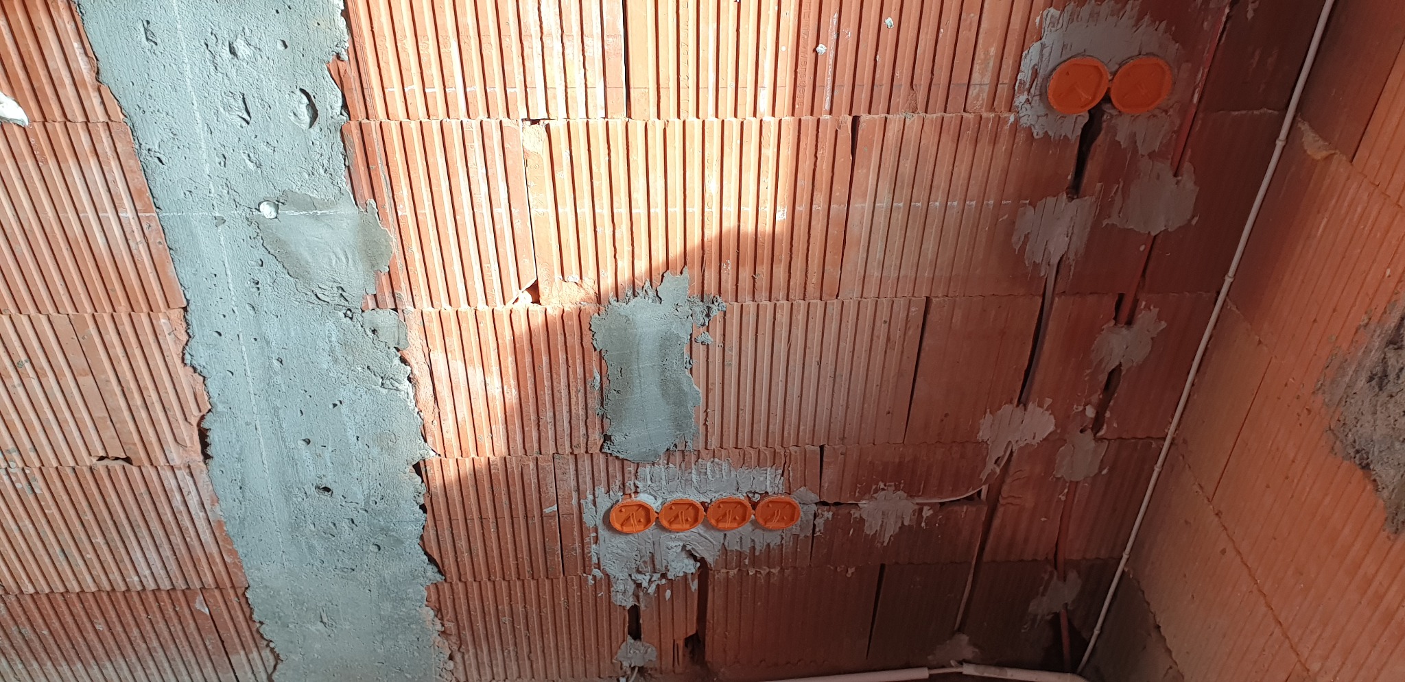 Ściana z cegły z wyżłobieniami wypełnionymi szarym cementem, widoczne pomarańczowe puszki elektryczne i biały kabel w peszlu.