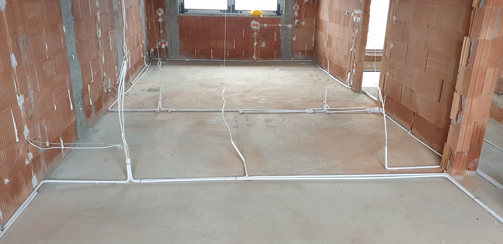 Instalacja elektryczna podtynkowa w surowym stanie, widoczne białe rurki PCV prowadzone po podłodze i ścianach z czerwonej cegły dziurawki, przygotowane pod wylewkę, żółty kask leży na parapecie...
