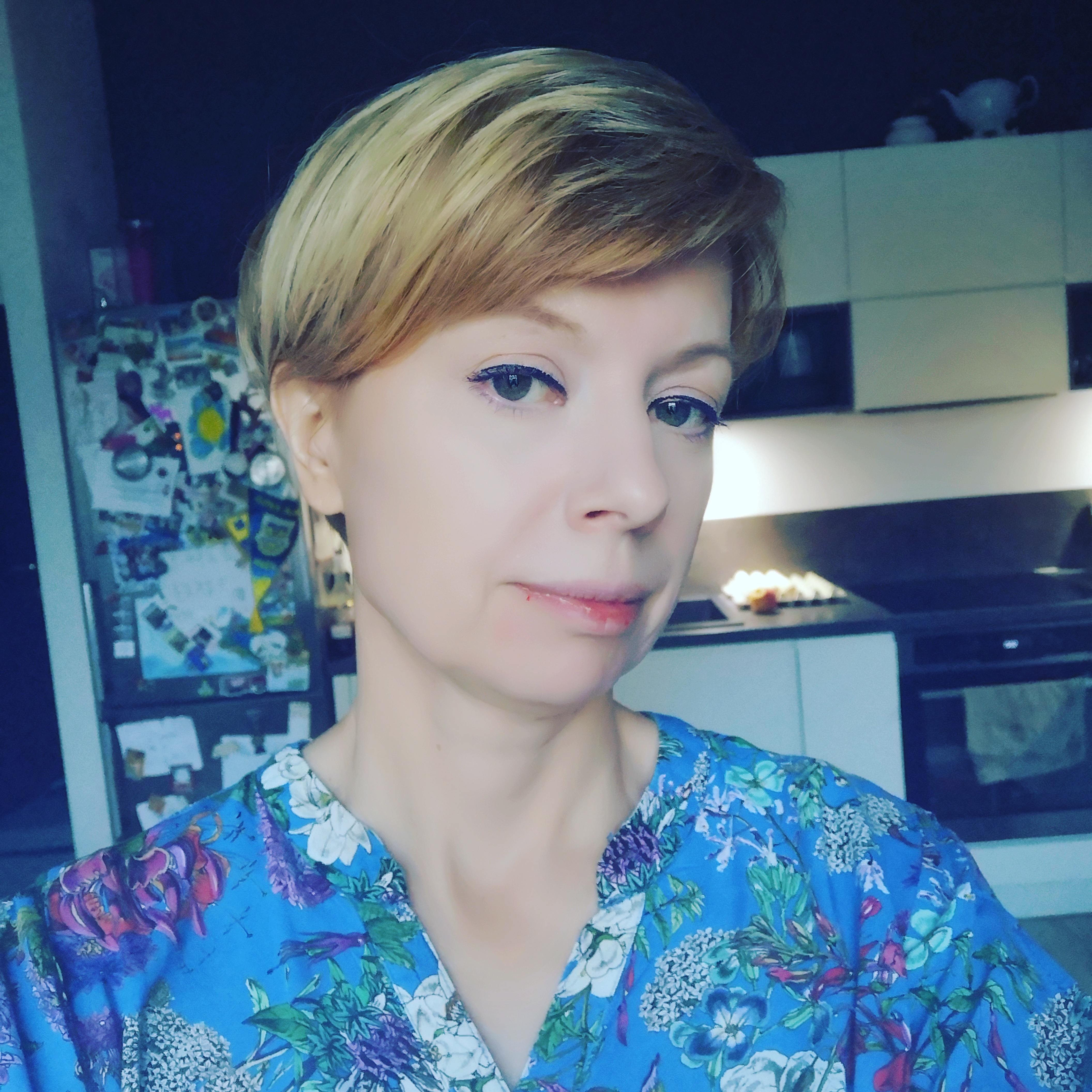 Portret nauczycielki angielskiego z krótkimi blond włosami w kwiecistej bluzce, na tle kuchni z lodówką ozdobioną magnesami.