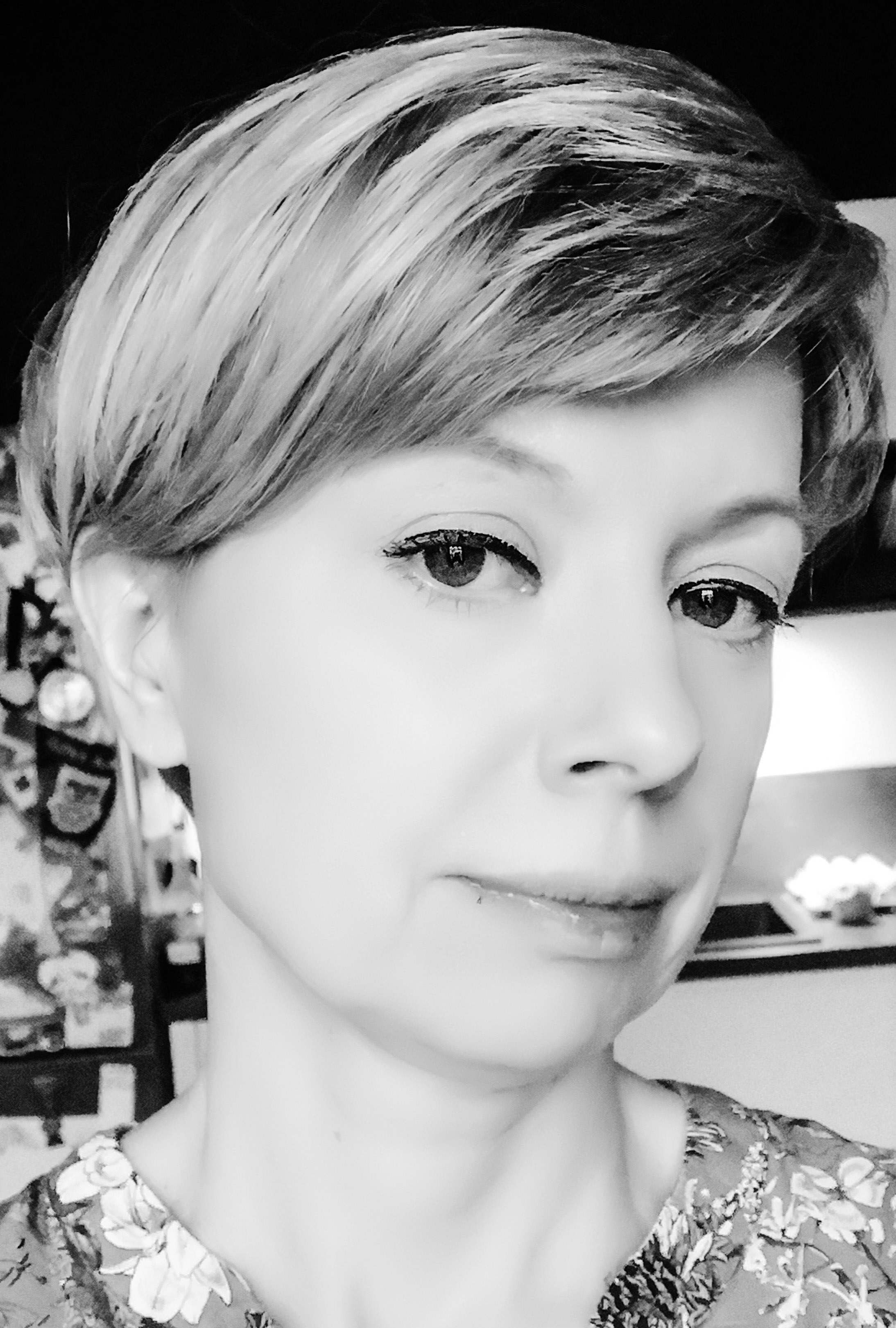 Portret kobiety z krótkimi blond włosami w czerni i bieli, z delikatnym uśmiechem, patrzącej wprost. W tle widoczne elementy wyposażenia wnętrza.