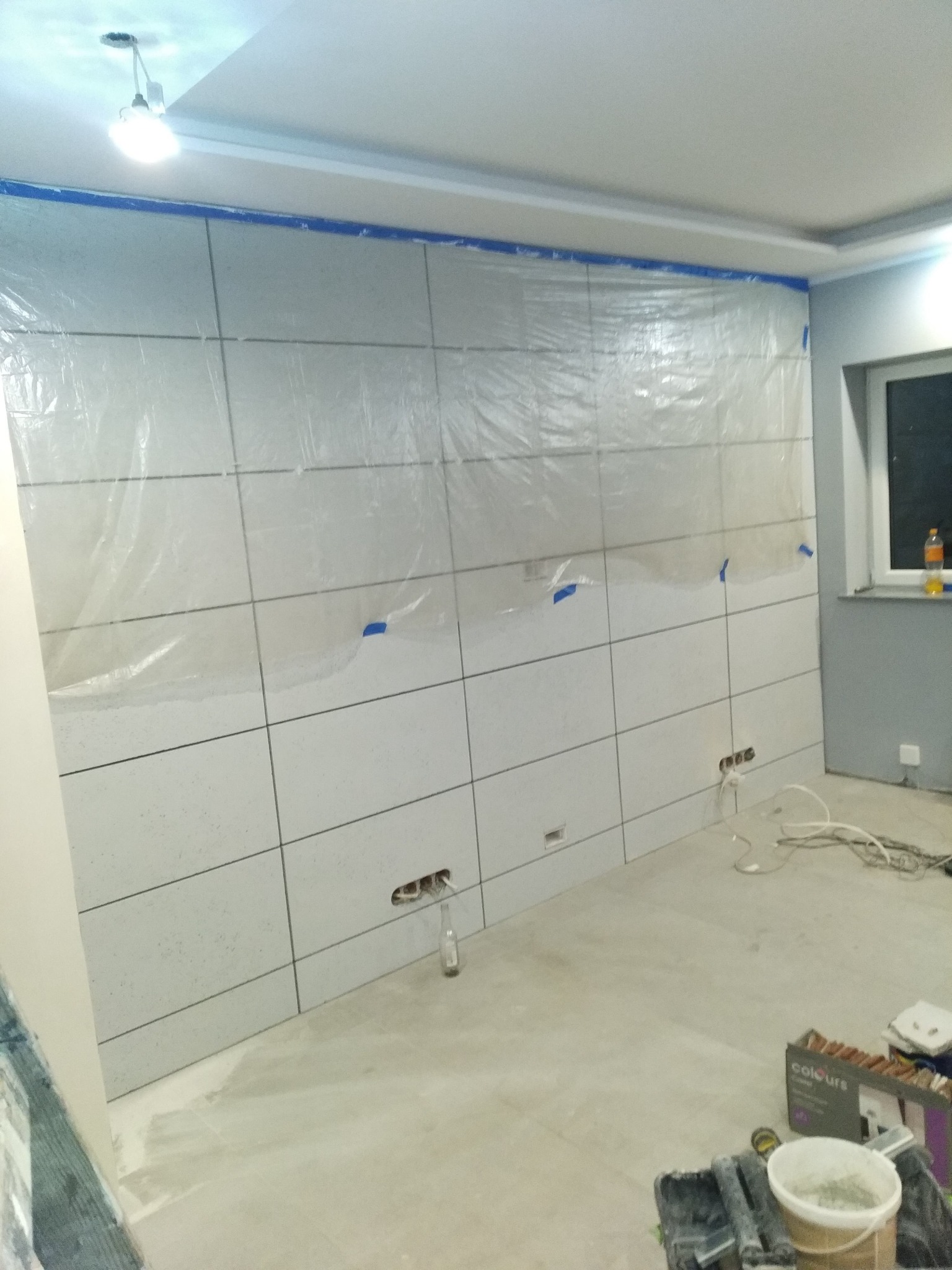 Ściana w trakcie układania płytek, zabezpieczona folią malarską, widoczne puszki elektryczne i akcesoria glazurnicze na podłodze.