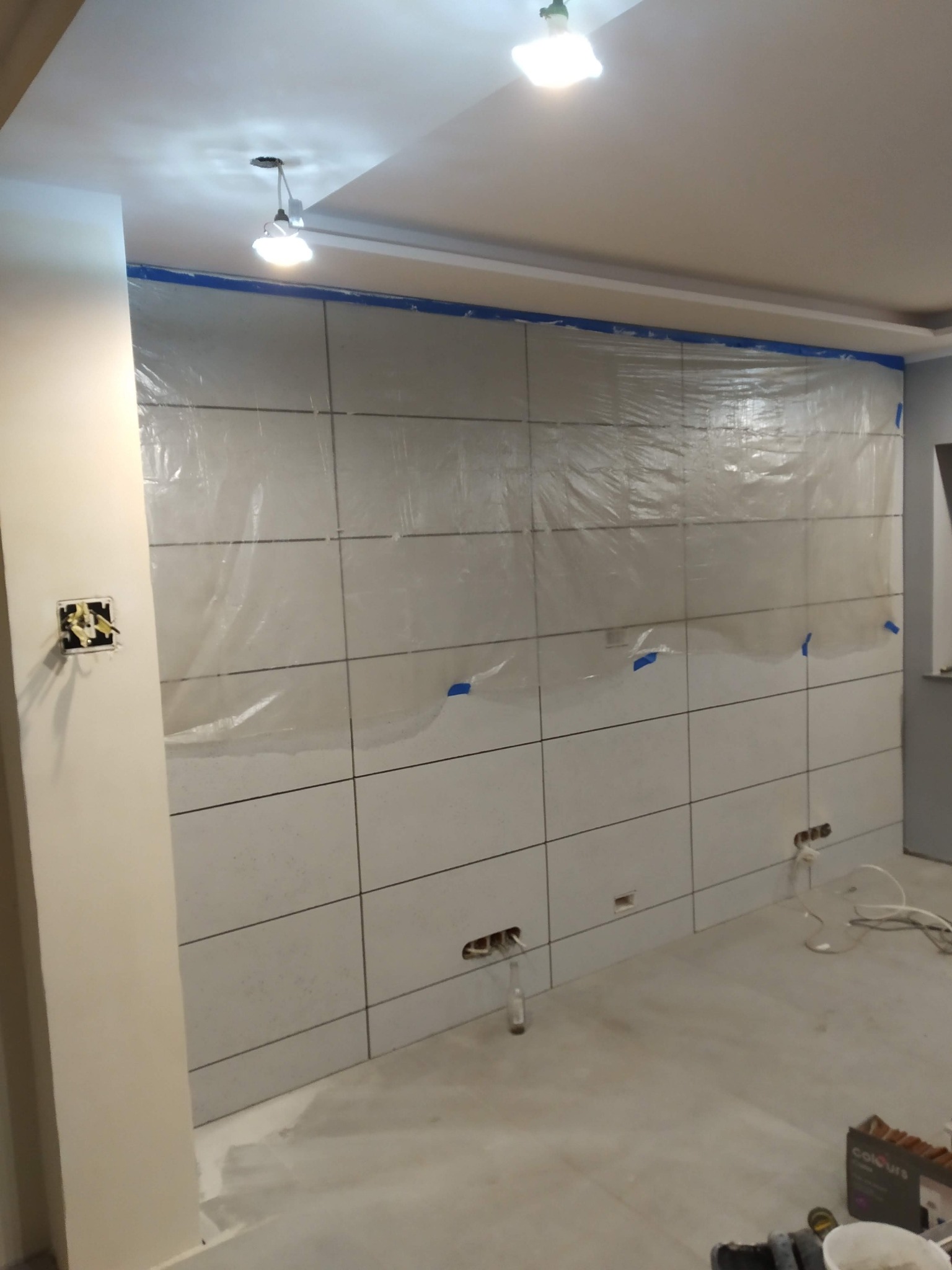 Ściana w trakcie układania jasnych, kwadratowych płytek, częściowo zabezpieczona folią malarską, widoczne puszki elektryczne i narzędzia na podłodze.