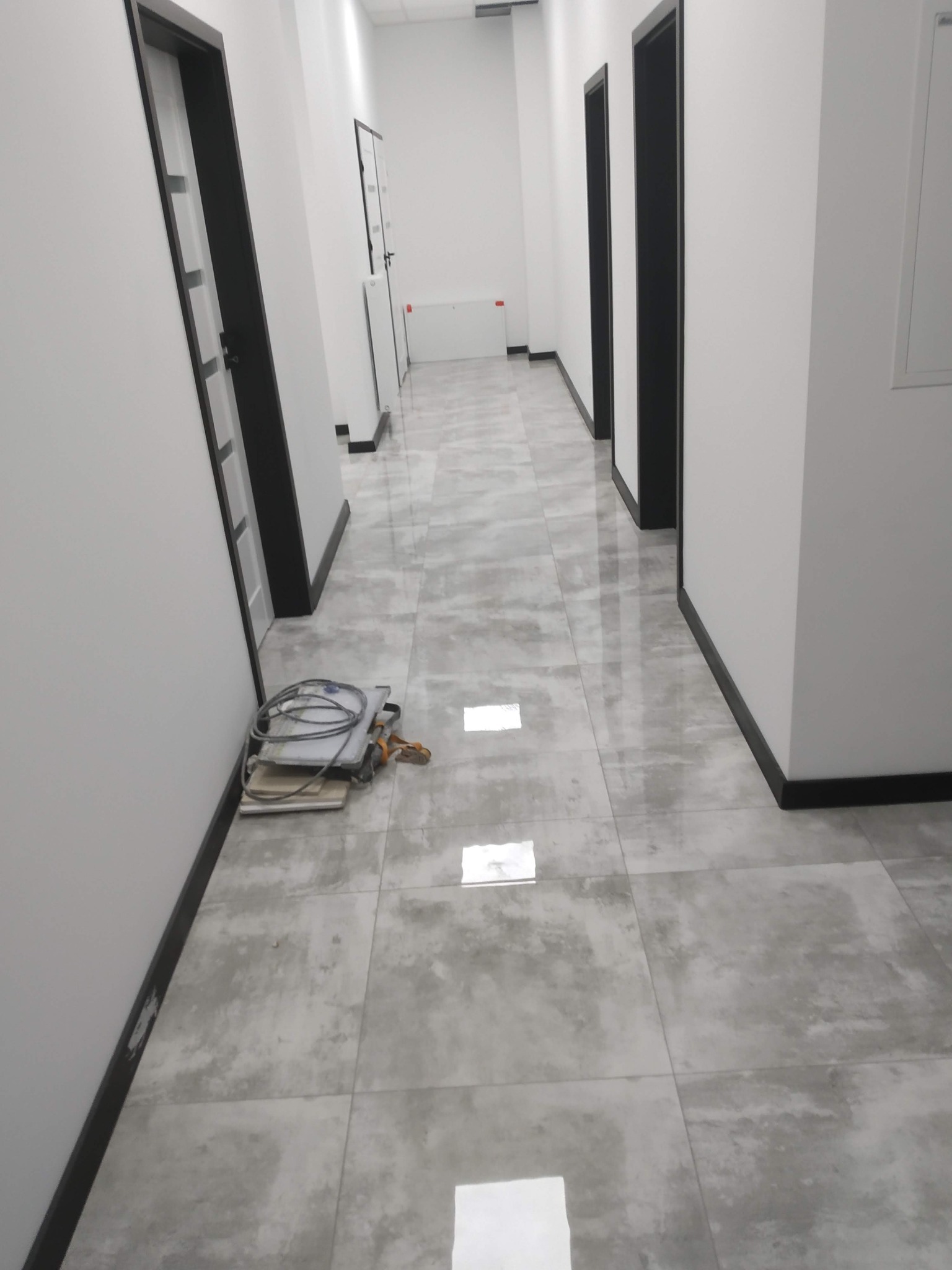 Korytarz z szarymi płytkami podłogowymi i białymi ścianami z czarnymi futrynami drzwi, na podłodze leżą narzędzia glazurnicze.