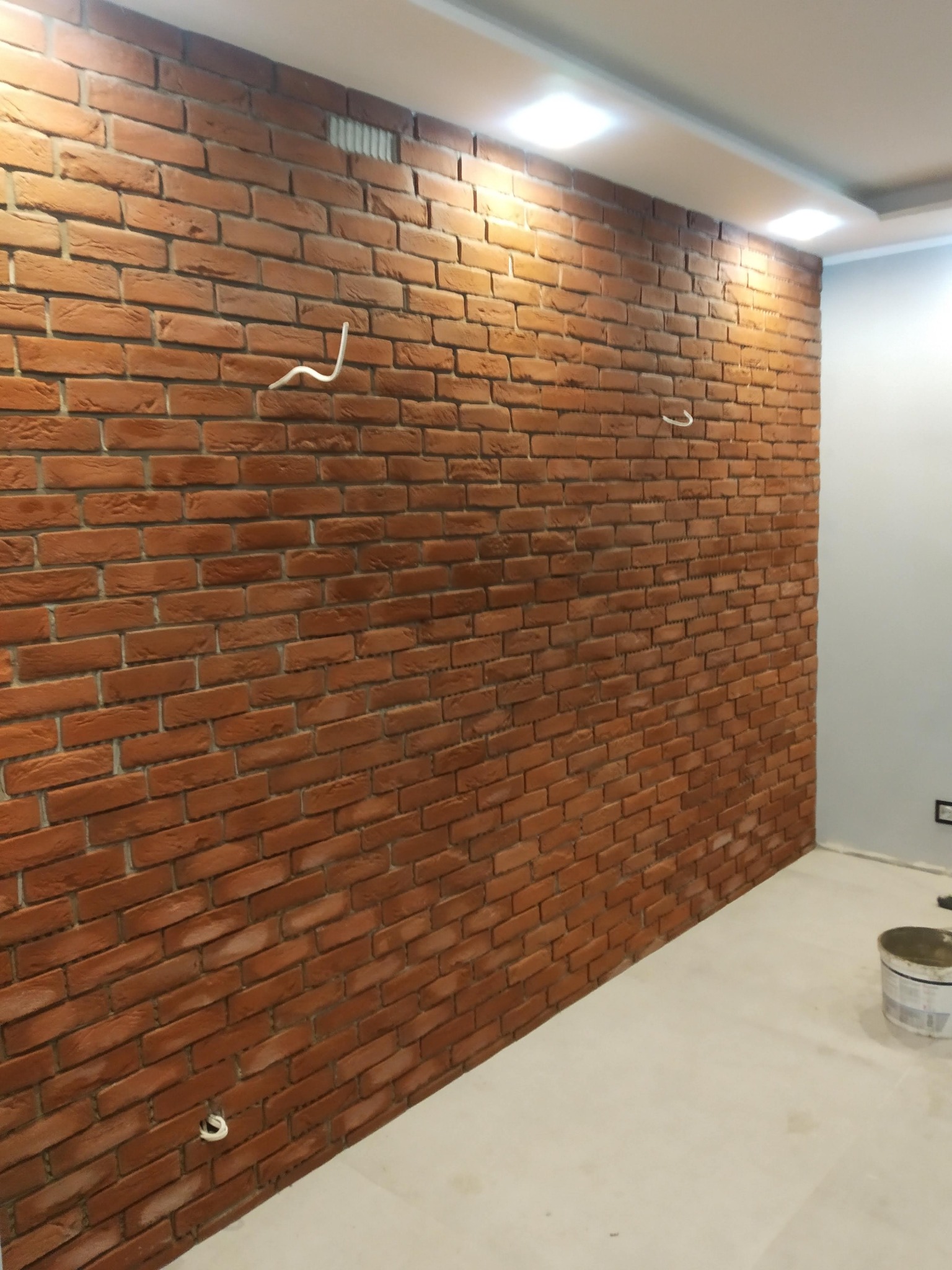 Ściana wyłożona cegłą klinkierową w odcieniach czerwieni i brązu, z widocznymi przewodami elektrycznymi. Na podłodze widoczne ślady po pracach wykończeniowych, obok ściany stoi wiadro z farbą.