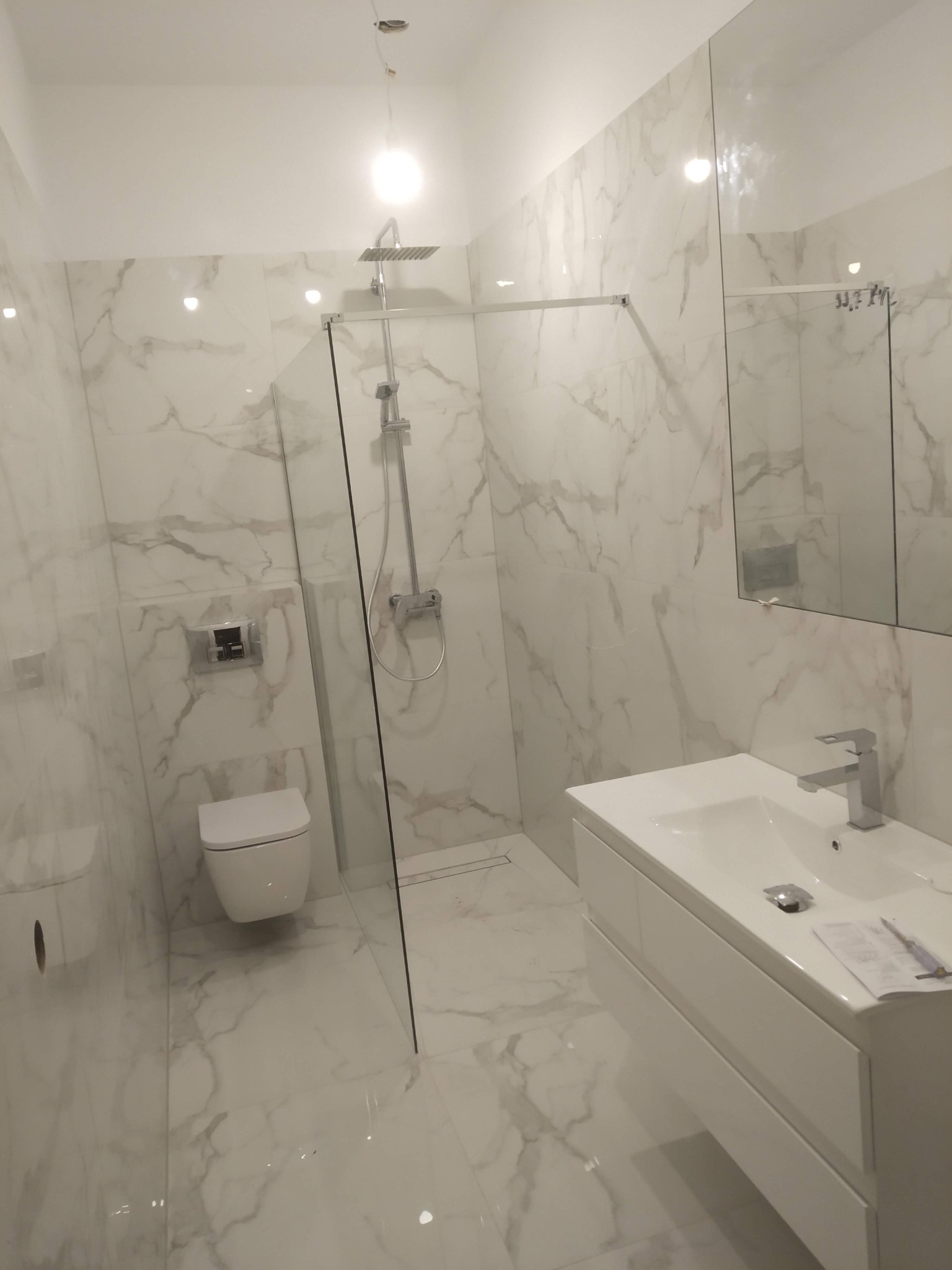 Nowoczesna łazienka z marmurowymi płytkami, przeszklonym prysznicem typu walk-in, wiszącą toaletą i białą szafką z umywalką, na której leżą dokumenty.