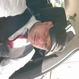 Mężczyzna w koszuli, kamizelce i krawacie, siedzący w samochodzie, prawdopodobnie w trakcie transportu. Widoczne zapięte pasy bezpieczeństwa.