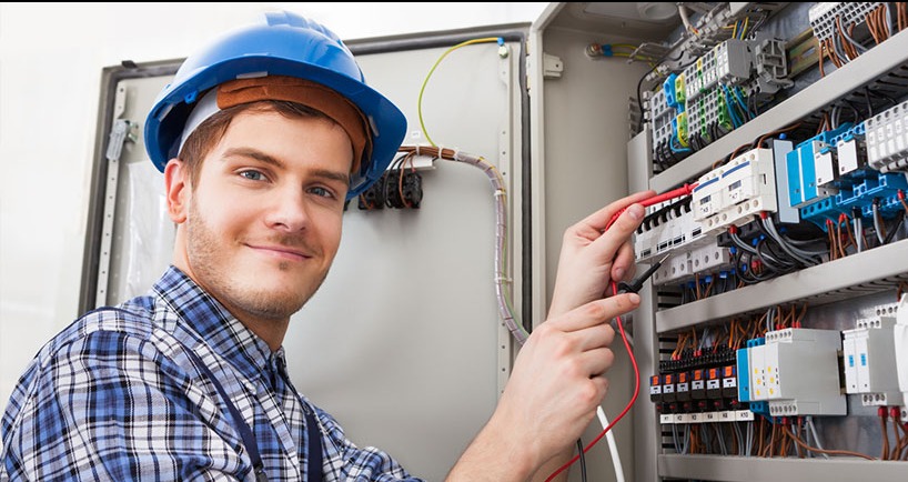 Elektryk w niebieskim kasku, z uśmiechem, mierzy napięcie w skrzynce elektrycznej. Widoczne przewody, bezpieczniki i miernik.