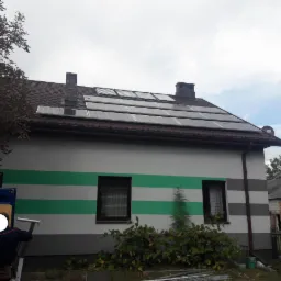 Instalacja paneli fotowoltaicznych na dachu domu jednorodzinnego z zielonymi pasami na elewacji. Widoczny fragment samochodu dostawczego i drabina.