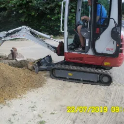 Mini koparka Takeuchi TB216 podczas pracy, operator w kabinie, dwaj mężczyźni obserwują wykop obok hydrantu, widoczne ślady gąsienic na piasku.