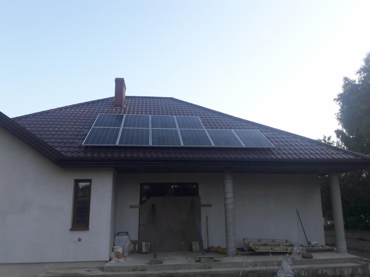 Panele słoneczne zamontowane na brązowym dachu domu jednorodzinnego z ceglanym kominem, widoczne materiały budowlane przy elewacji.