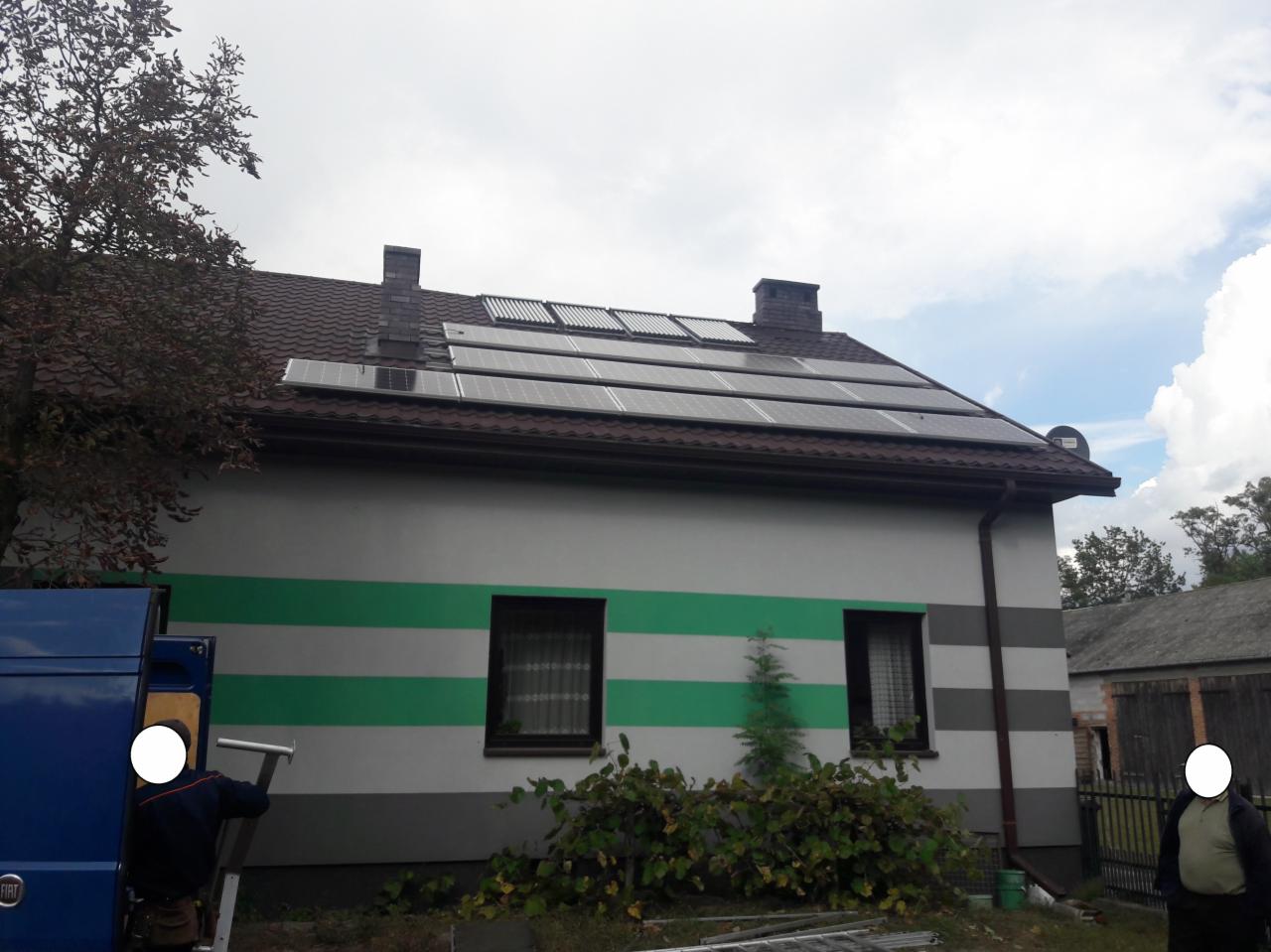 Instalacja paneli fotowoltaicznych na dachu domu jednorodzinnego z zielonymi pasami na elewacji. Widoczny fragment samochodu dostawczego i drabina.