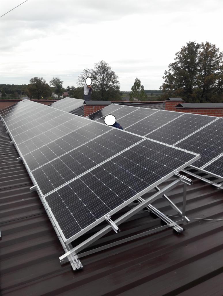 Instalacja paneli fotowoltaicznych na brązowym dachu z blachy trapezowej. Widoczne aluminiowe konstrukcje wsporcze i osoby pracujące na dachu. Pochmurne niebo w tle.