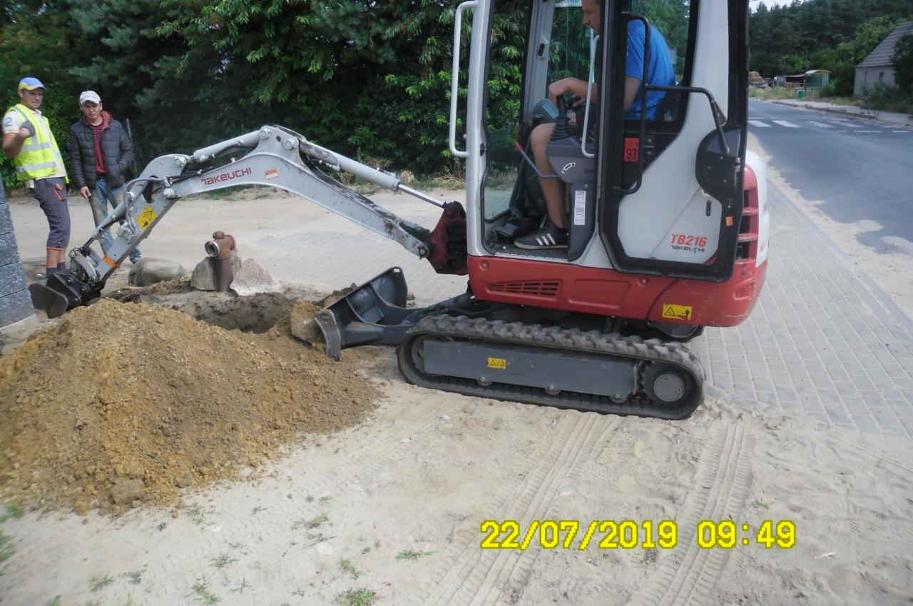 Mini koparka Takeuchi TB216 podczas pracy, operator w kabinie, dwaj mężczyźni obserwują wykop obok hydrantu, widoczne ślady gąsienic na piasku.