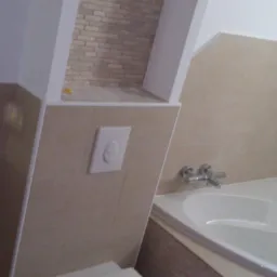 Nowoczesna łazienka z narożną wanną, beżowymi płytkami i ukrytą spłuczką w ścianie, dodatkowo widoczna dekoracyjna wnęka wyłożona płytkami imitującymi cegłę.