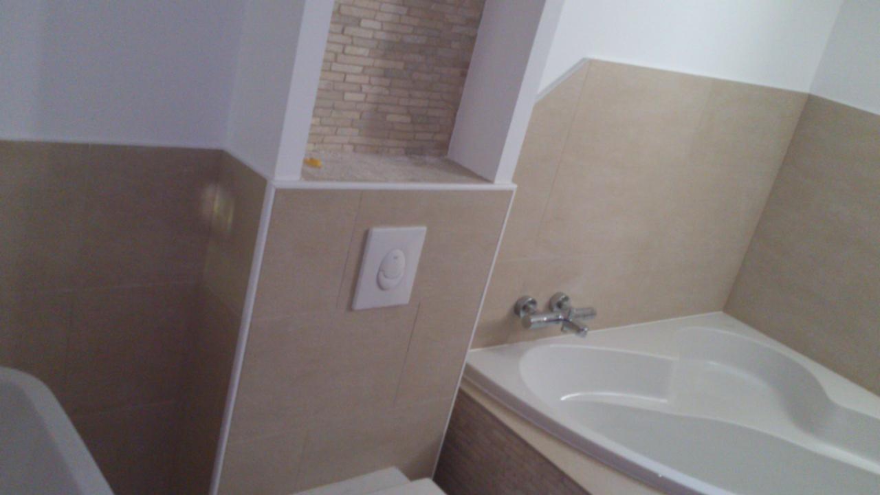 Nowoczesna łazienka z narożną wanną, beżowymi płytkami i ukrytą spłuczką w ścianie, dodatkowo widoczna dekoracyjna wnęka wyłożona płytkami imitującymi cegłę.