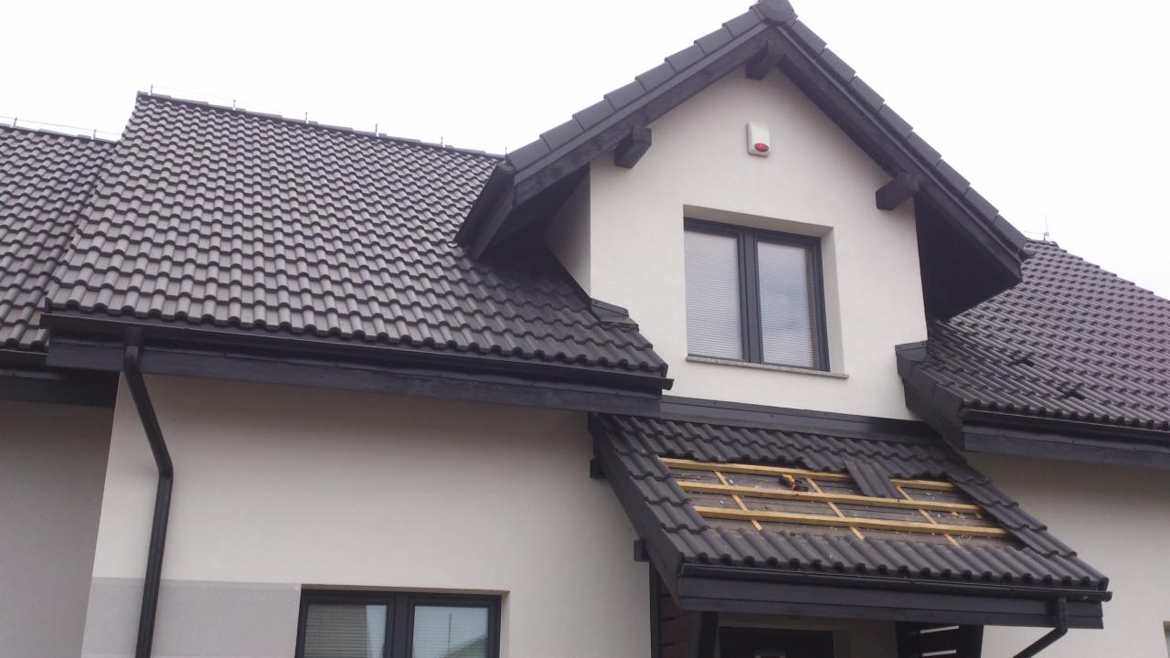 Fragment dachu dwuspadowego z czarną dachówką ceramiczną w trakcie naprawy, widoczne łaty pod dachówkami, biała elewacja budynku z czarnymi oknami i rynnami.