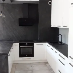 Nowoczesna kuchnia z białymi szafkami i czarnym blatem, czarna ściana nad blatem, oświetlenie punktowe sufitowe, minimalistyczny design.