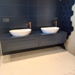 Dwie białe umywalki nablatowe z miedzianymi bateriami na szafce w kolorze ciemnego granatu, na tle ściany z ciemnogranatowych płytek, częściowo wykończonej heksagonalnymi płytkami, widoczne...