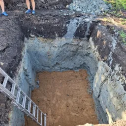 Głęboki wykop w ziemi z widocznymi warstwami gleby, metalowa drabina oparta o ścianę wykopu, w tle pryzma ziemi i fragmenty sylwetek dwóch osób.