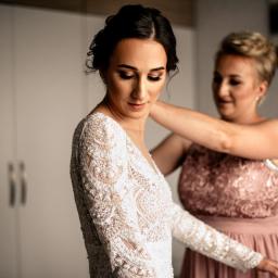 Kawka Art - Panna młoda w koronkowej sukni z długim rękawem, poprawiana przez kobietę w różowej sukience, przygotowania do ślubu.
