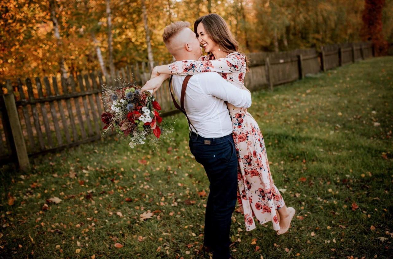 Uśmiechnięty mężczyzna w białej koszuli i granatowych spodniach z szelkami podnosi kobietę w kwiecistej sukience na tle jesiennego krajobrazu i drewnianego płotu; bukiet kwiatów w dłoni.