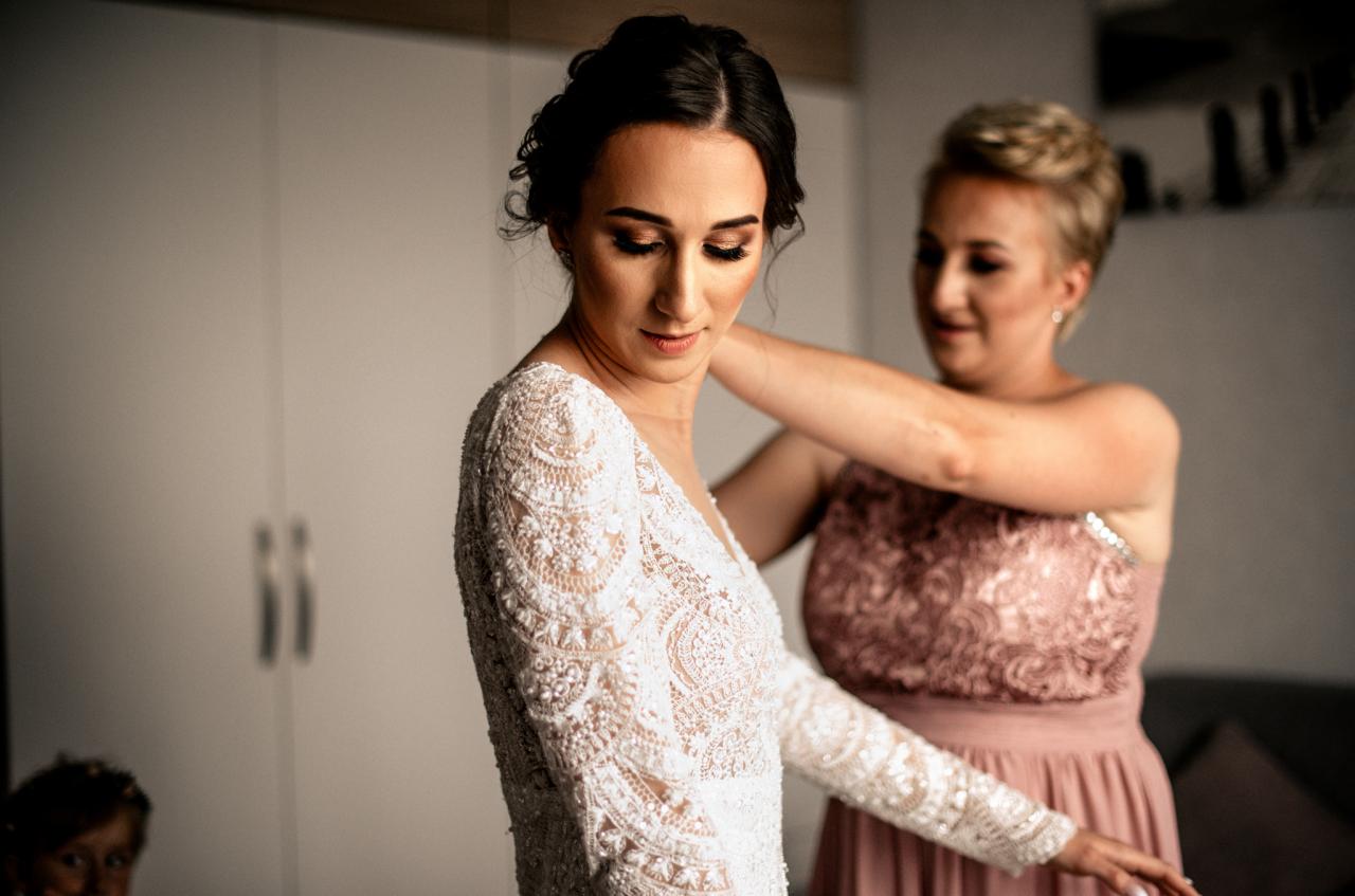 Panna młoda w koronkowej sukni z długim rękawem, poprawiana przez kobietę w różowej sukience, przygotowania do ślubu.