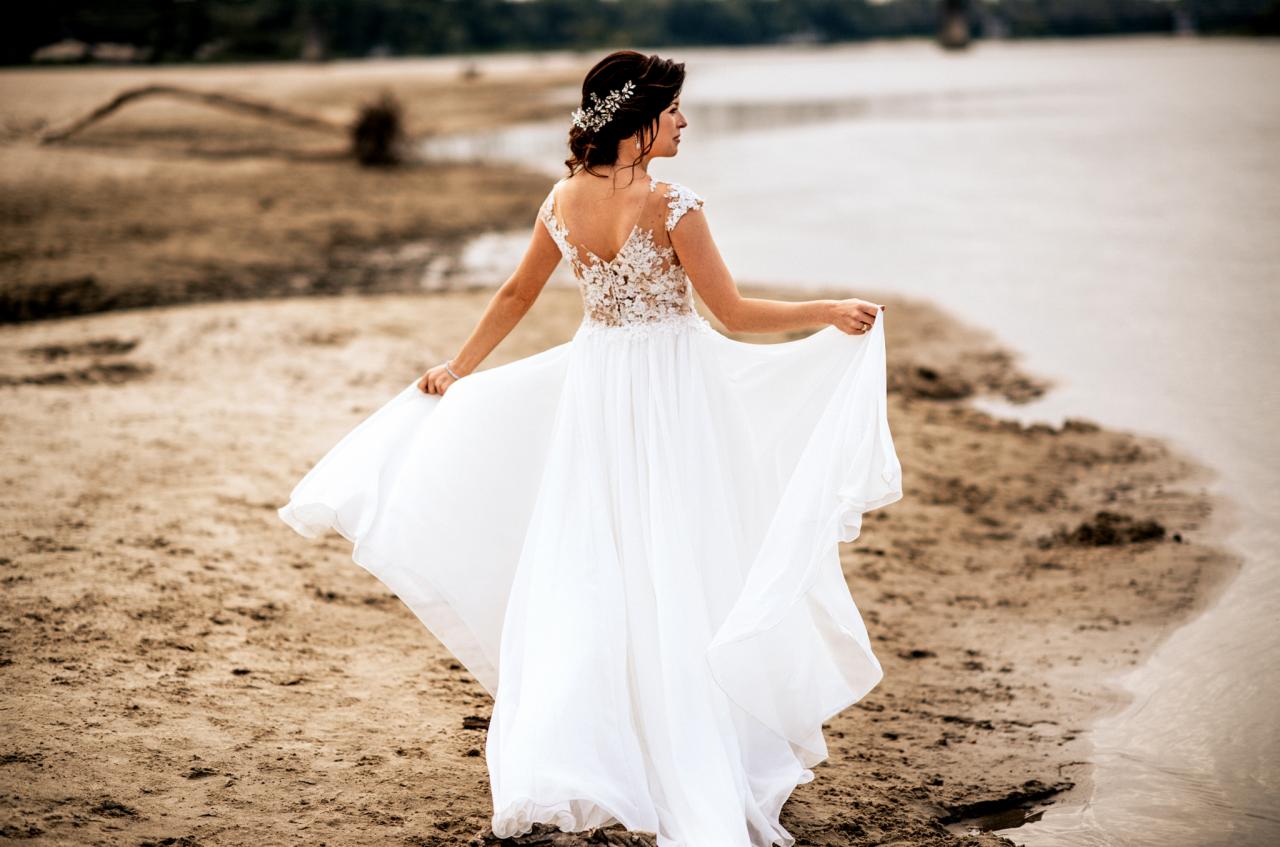 Panna młoda w białej sukni z koronkową górą i ozdobą we włosach, stojąca tyłem na piaszczystej plaży przy brzegu wody.