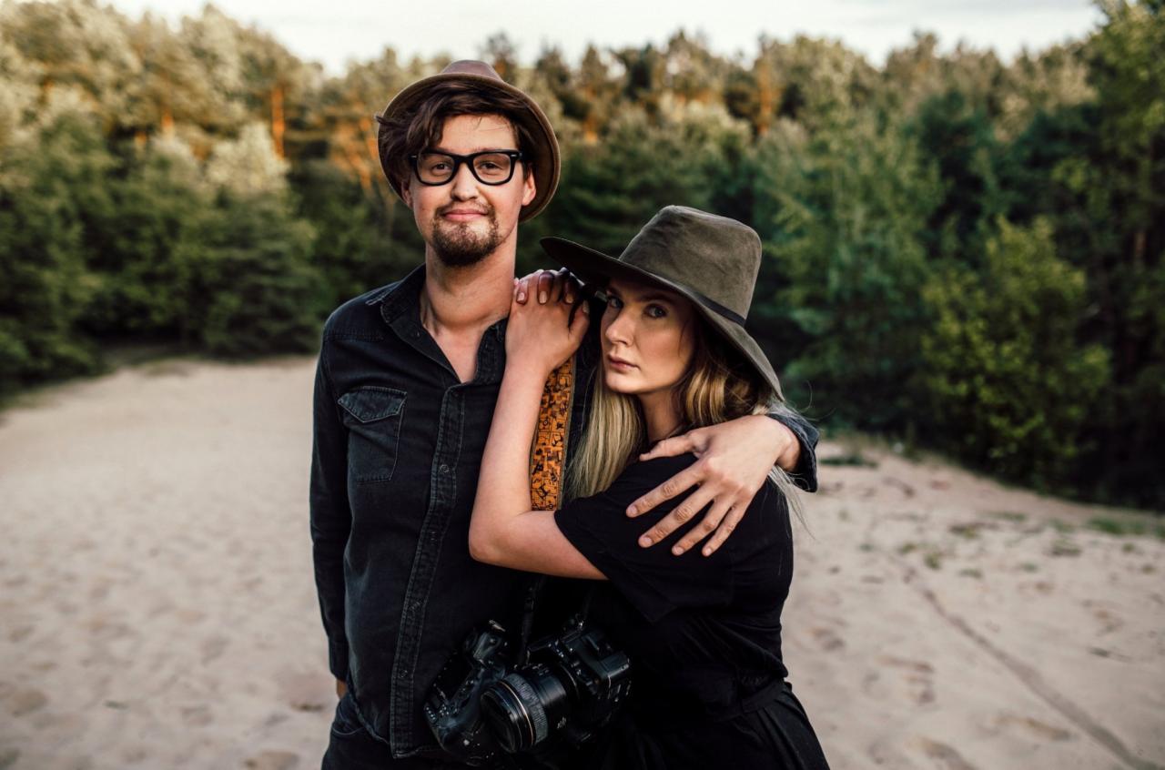 Para fotografów w plenerze: mężczyzna w okularach i kapeluszu, kobieta z aparatem, oboje w ciemnych ubraniach na tle piaszczystego wzgórza i lasu.