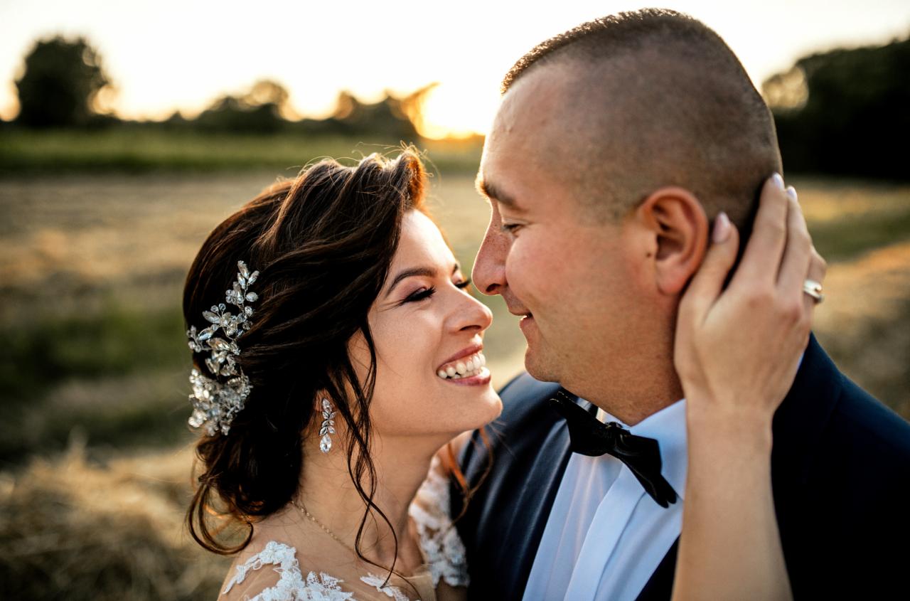 Uśmiechnięta para młoda, panna młoda z ozdobą we włosach i kolczykami, pan młody w muchą, delikatne światło zachodzącego słońca w tle.