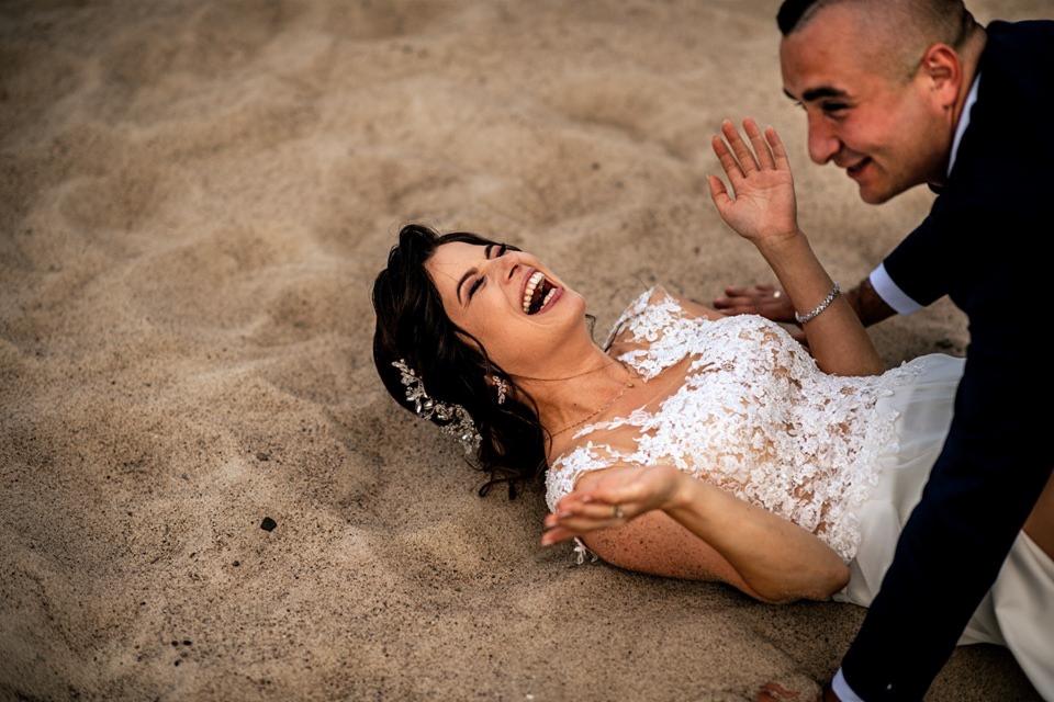Panna młoda w koronkowej sukni ślubnej leży na piasku, śmiejąc się w stronę pana młodego z podgolonymi włosami, który wyciąga do niej rękę.