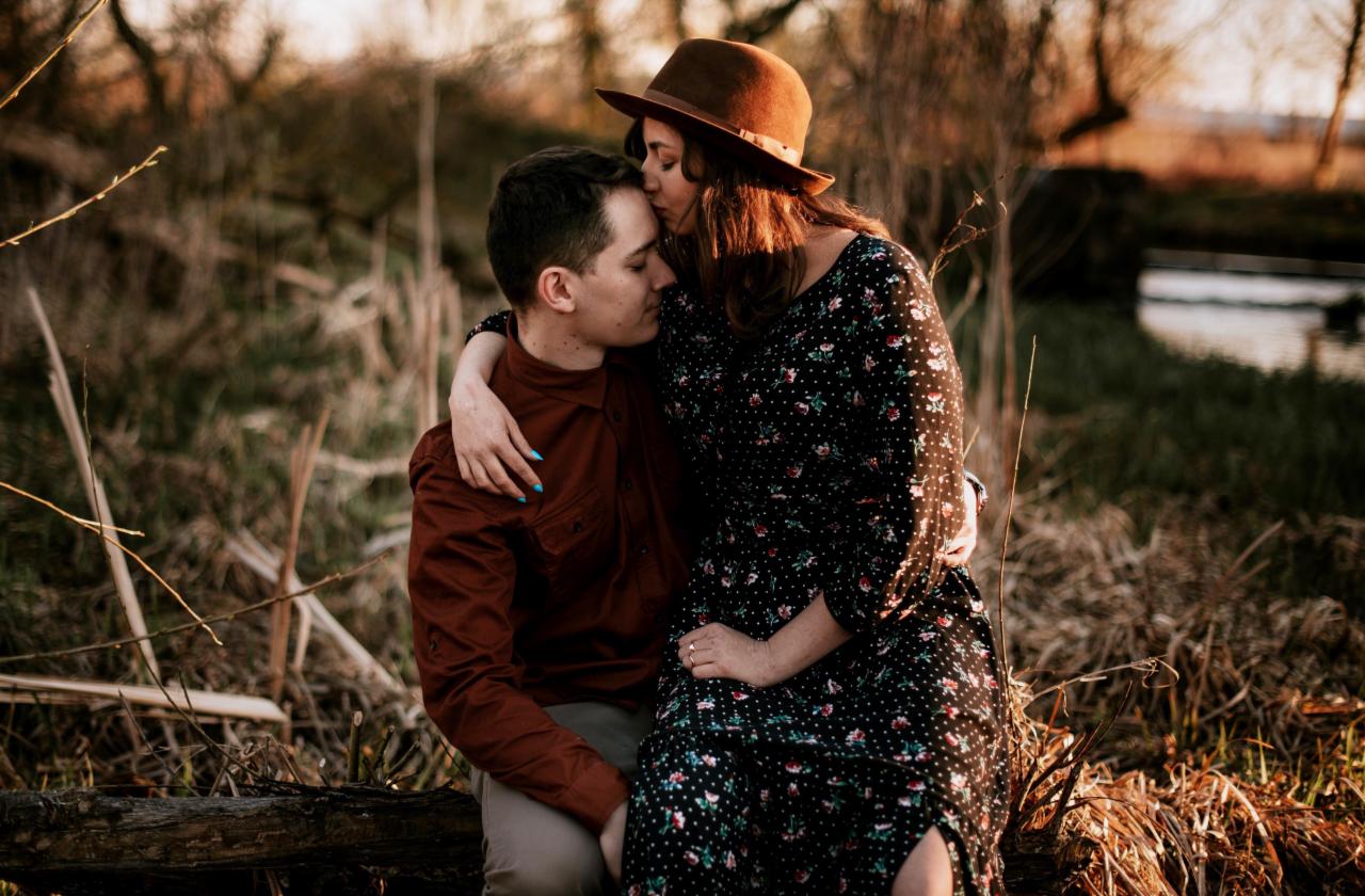 Zakochana para siedząca na pniu drzewa w plenerze, kobieta w kapeluszu całuje mężczyznę w czoło, w tle trawa i drzewa.