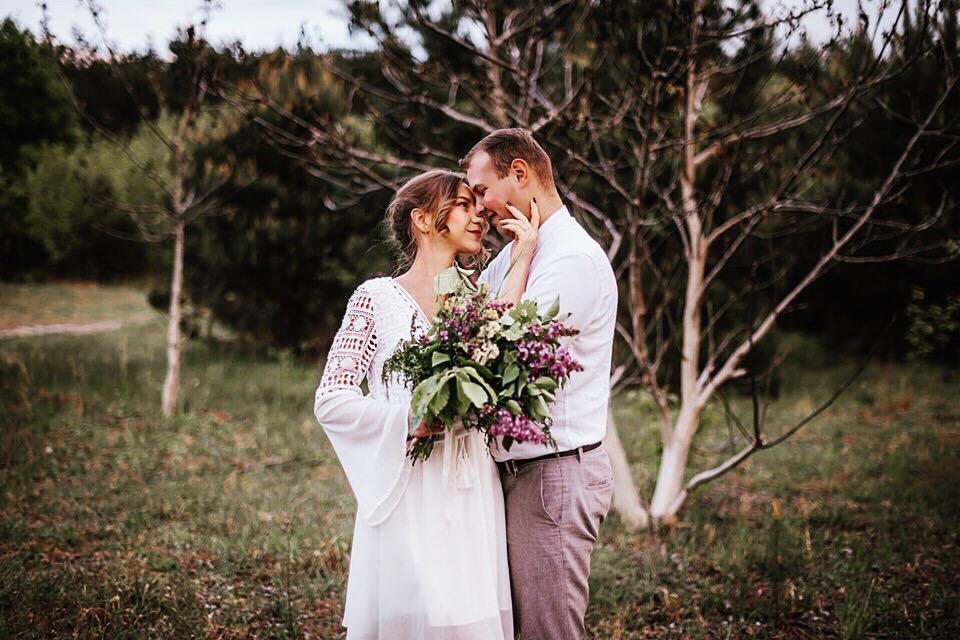 Zakochana para w stylu boho, trzymająca bukiet polnych kwiatów, sfotografowana w plenerze w Warszawie, z drzewami w tle.