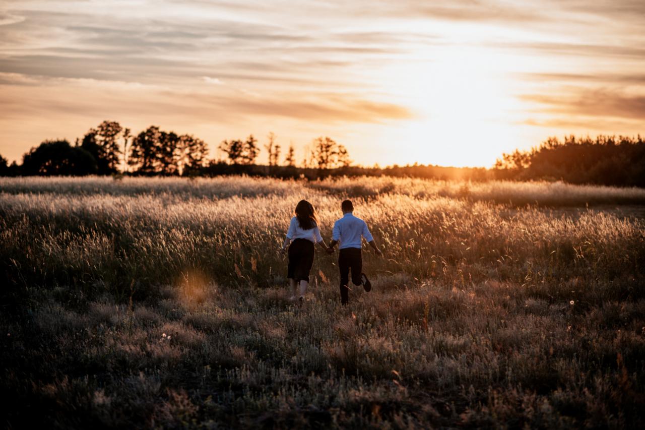 Para trzymająca się za ręce biegnie przez pole traw w kierunku zachodzącego słońca, sylwetki drzew w tle.