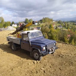 Niebieski Land Rover Defender z wiertnicą zamontowaną na pace, stojący na placu budowy z widokiem na pagórkowaty krajobraz i zabudowania.