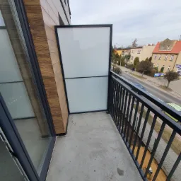 Balkon z czarną, metalową balustradą i szklaną osłoną, widok na ulicę z budynkami i przejeżdżającymi samochodami.