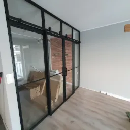 Przeszklona ścianka działowa z czarnymi profilami, przesuwne drzwi z widokiem na ceglaną ścianę, wnętrze w trakcie wykańczania, kartony na podłodze.