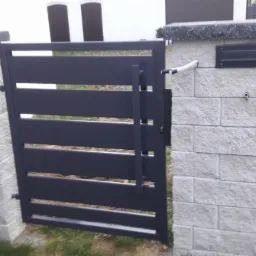 Nowoczesna, pozioma brama wjazdowa z ciemnoszarego metalu, zamontowana między dwoma słupkami z jasnoszarego kamienia, z widoczną skrzynką na listy na jednym ze słupków.