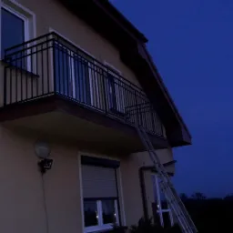Czarna balustrada balkonowa z pionowymi prętami, zamontowana na budynku z beżową elewacją, widoczna drabina oparta o balkon w wieczornym oświetleniu.