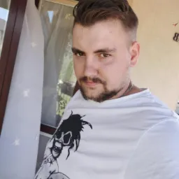 Mężczyzna z brodą w białej koszulce z nadrukiem, stojący wewnątrz, obok okna z białymi firankami i brązową ramą.