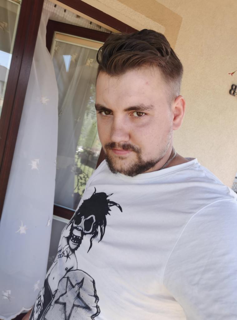 Mężczyzna z brodą w białej koszulce z nadrukiem, stojący wewnątrz, obok okna z białymi firankami i brązową ramą.