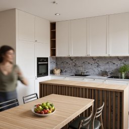 JELCONSULTING SP. Z O.O. - Jasna kuchnia z wyspą i drewnianym stołem, na którym leżą owoce. W tle rozmyta sylwetka kobiety. Minimalistyczny design z szarym blatem i kamienną ścianą.