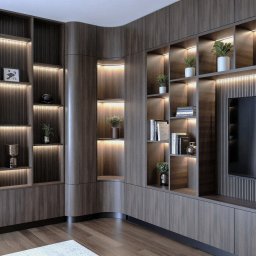 JELCONSULTING SP. Z O.O. - Elegancka zabudowa meblowa z ciemnego drewna z ryflowanymi panelami, zakrzywionym narożnikiem i podświetlanymi półkami. Zintegrowany telewizor i dekoracje.