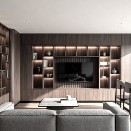 JELCONSULTING SP. Z O.O. - Nowoczesny salon z zabudową meblową z ciemnego drewna, podświetlanymi półkami, zintegrowanym telewizorem i minimalistycznym wystrojem. Widoczna szara sofa i stół jadalniany.