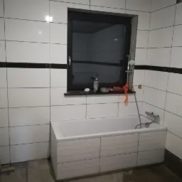 Białe, prostokątne płytki ścienne z czarnym pasem dekoracyjnym, okno z czarną ramą nad wanną z panelami o falistym wzorze, widoczny fragment drabiny malarskiej.